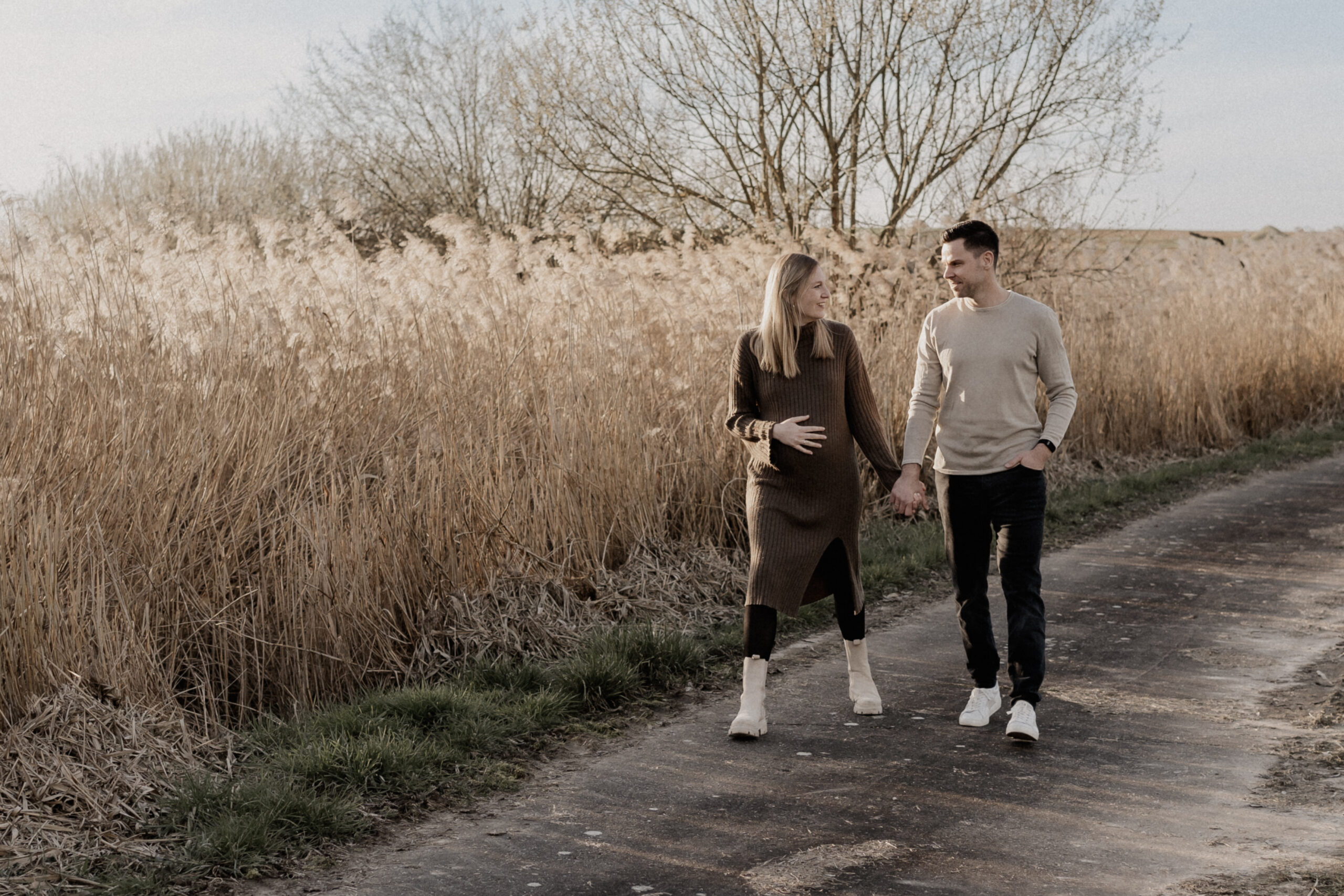 Babybauchshooting mit einem Paar. Beide laufen Hand in Hand einen Weg entlang.